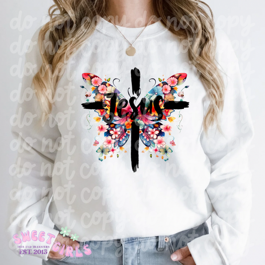 A woman wearing a white sweatshirt featuring a vibrant design of a cross with the word “Jesus” overlaying it, surrounded by a colorful butterfly made up of various flowers. The design is bold and eye-catching, symbolizing faith and transformation. The woman is also wearing a simple necklace and has her hands in her jeans pockets. The background is plain, emphasizing the detailed artwork on the sweatshirt.